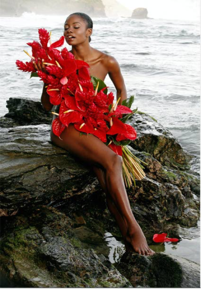 A woman with dark skin sits on rocks by the ocean, holding a large bouquet of vibrant red flowers, creating an artistic seaside scene.