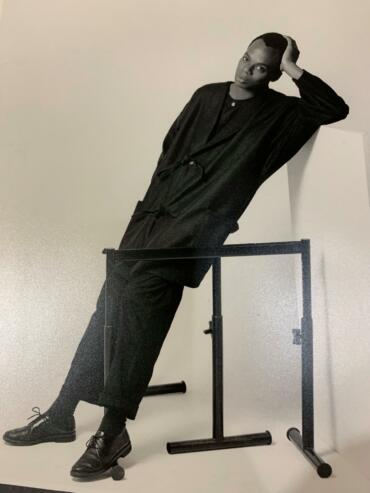 Black-and-white portrait of a woman in a loose suit, leaning on a barre against a plain wall.