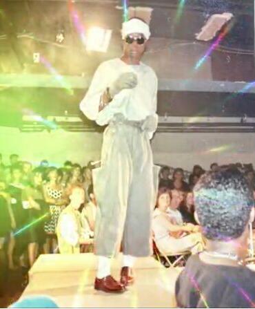 Person on a stage wearing stylish gray pants, a hat, sunglasses, and gloves at a fashion event.