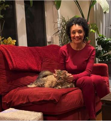 Denise wearing a red sweater and pants, sitting on a red sofat with her cat at home.