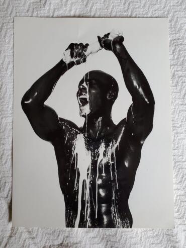 Black-and-white photo of a male figure with dark skin, pouring a white liquid over his head and body.
