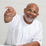 An older man with glasses, a beard, and a white perforated shirt, smiling and holding out a ring.
