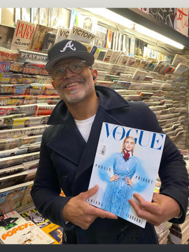 Man holding a Vogue Italia magazine in a bookstore, dressed in a dark coat and glasses.