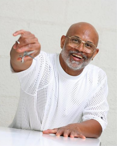 An older man with glasses, a beard, and a white perforated shirt, smiling and holding out a ring.