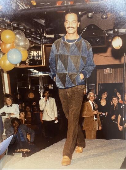 Man walking on runway at a party, wearing a geometric-patterned sweater and brown pants.