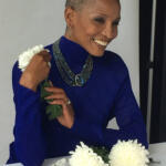 Elegant woman with a shaven head, wearing a vibrant blue top, jewelry, and holding white chrysanthemums.