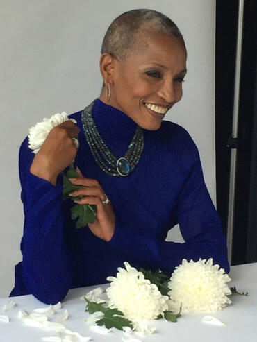 Elegant woman with a shaven head, wearing a vibrant blue top, jewelry, and holding white chrysanthemums.