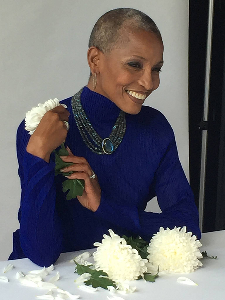 Elegant woman with a shaven head, wearing a vibrant blue top, jewelry, and holding white chrysanthemums.