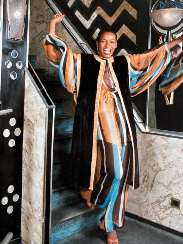 Woman in vibrant striped jumpsuit and long coat smiling joyfully near mirror and staircase.