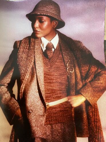 Woman in a houndstooth hat, layered brown textured coat and sweater, with white shirt and tie.