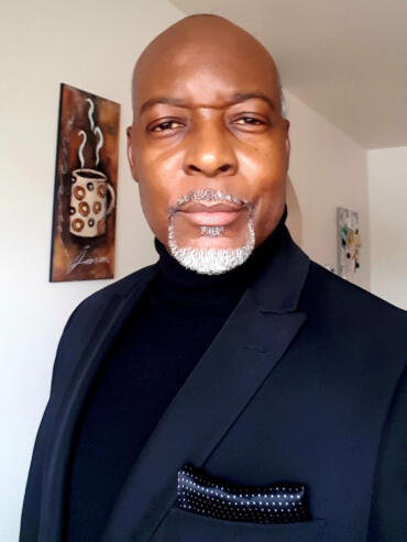 Elegant man in a black blazer and turtleneck, sporting a neatly groomed gray beard.