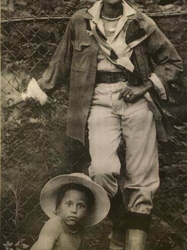 Stylish woman with head wrap and layered clothing, standing outdoors with a young girl in a straw hat, suggesting a vintage fashion scene.