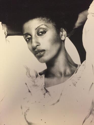 Black-and-white portrait of a woman with confident expression, wearing a stylish top, and resting her arm on her head.