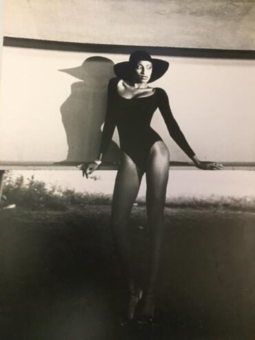 Black-and-white fashion photo of a woman in a wide-brimmed hat and bodysuit, posing outdoors.