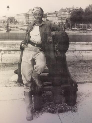 A woman in vintage fashion, sitting on a park bench with hands on her lap, wearing a headscarf, leather jacket, and high-waisted trousers, captured in an urban setting.