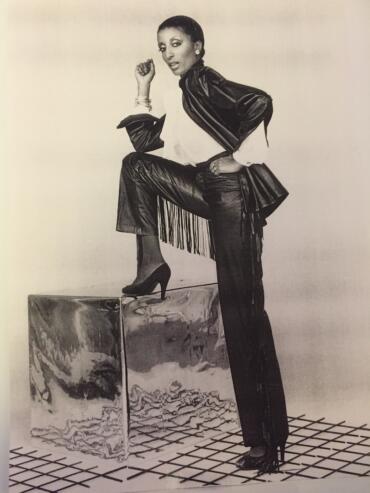 A woman in leather pants and jacket, posed confidently on a reflective cube, showcasing 1980s fashion style.