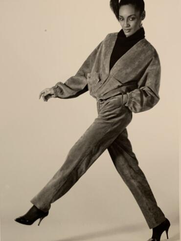 Black and white photo of Denise taking a wide step, wearing a suede jacket and matching ankle length straight leg pants, her hair in a ponytail.