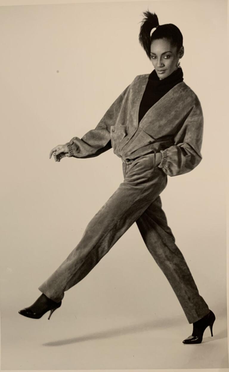 Black and white photo of Denise taking a wide step, wearing a suede jacket and matching ankle length straight leg pants, her hair in a ponytail.