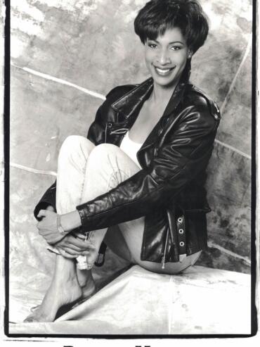 Black-and-white portrait of Donna Holgate with a leather jacket, sitting barefoot, smiling in a studio setting.