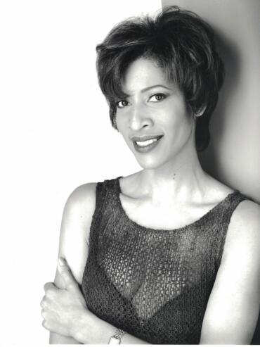Black and white portrait of a woman with short, tousled hair, wearing a sleeveless, textured top, smiling confidently.