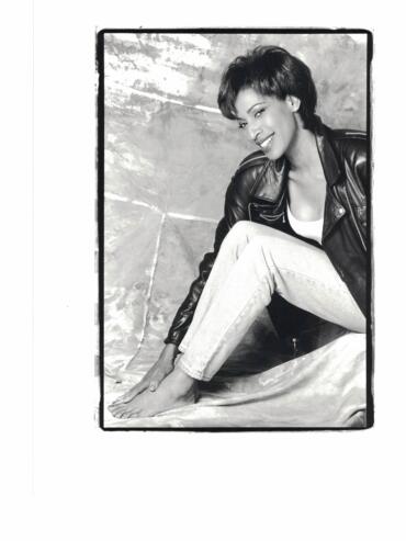 Black-and-white portrait of a woman sitting barefoot, wearing a leather jacket and smiling.
