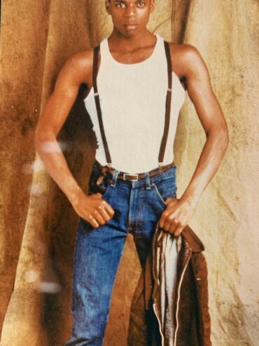 Androgynous model in jeans, suspenders, and a sleeveless top against a textured backdrop.