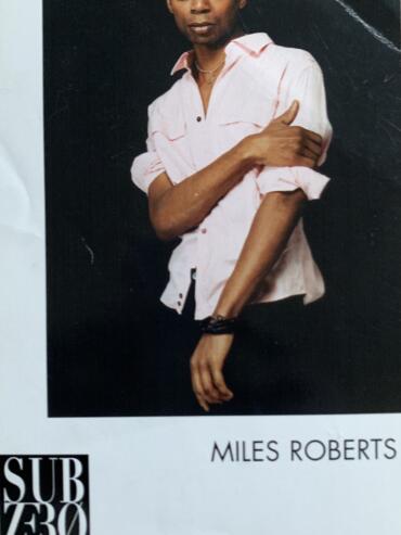 Portrait of a man in a pink shirt with rolled-up sleeves, posing against a dark background.