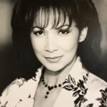 Black-and-white portrait of an elegant woman with mid-length hair, wearing a floral blouse and a matching beaded necklace.