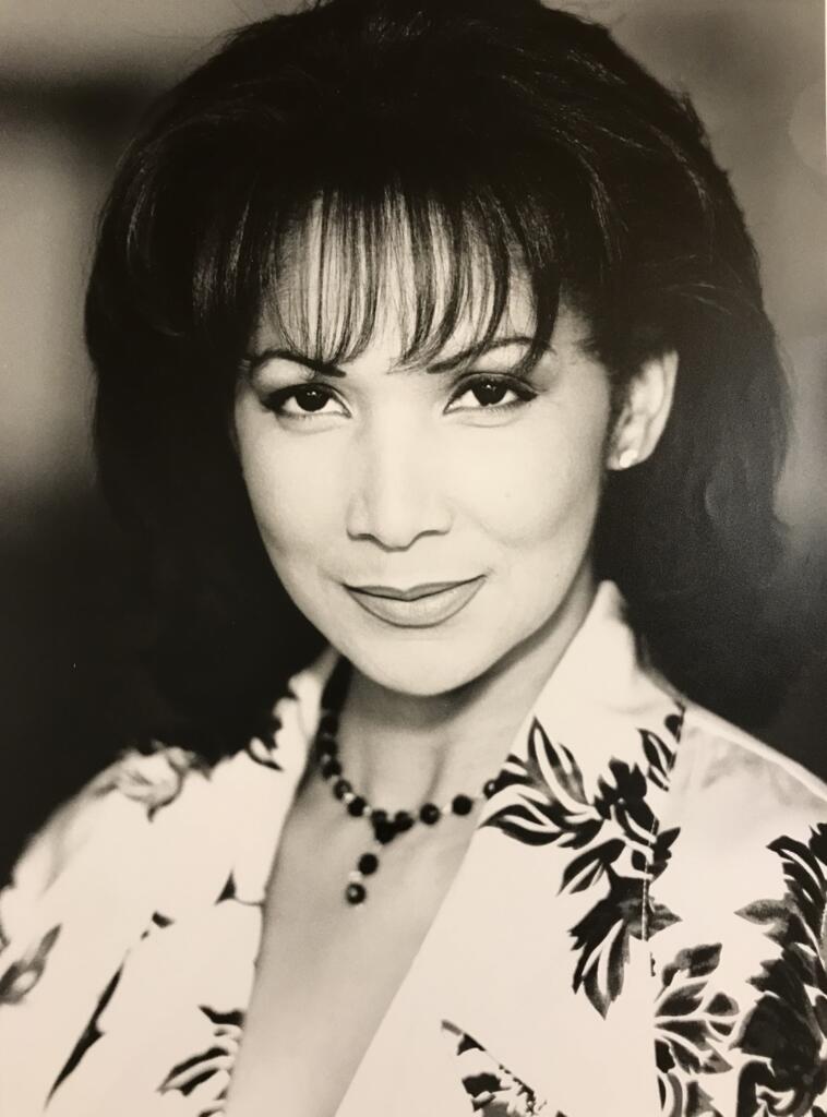Black-and-white portrait of an elegant woman with mid-length hair, wearing a floral blouse and a matching beaded necklace.
