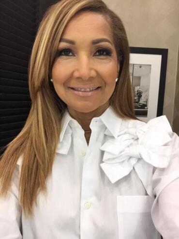 Woman in a white blouse with a ruffled shoulder detail, smiling softly.