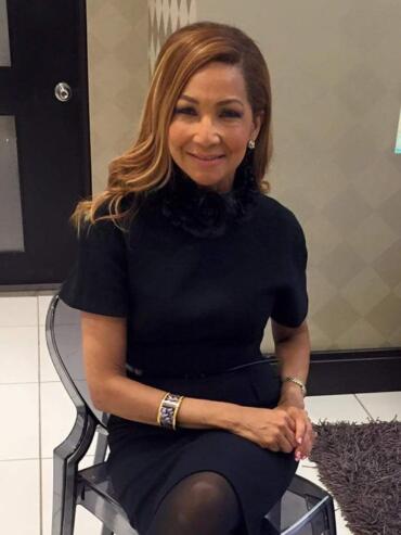 Elegant woman in a black dress featuring a ruffled collar, seated indoors with a welcoming smile.