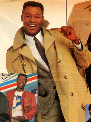 Man smiling in a beige trench coat and patterned tie.