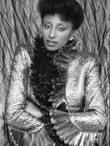Elegant woman in ornate jacket with ruffled collar and textured fabric, posed confidently.