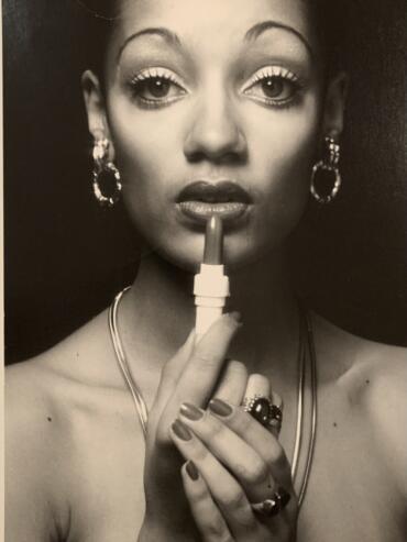 Black and white photo, head and shoulders, of Denise holding a lipstick with both hands, looking directly into the camera.