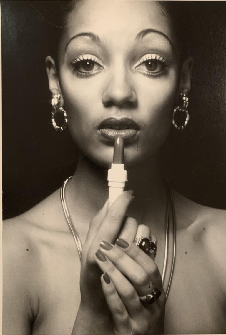 Black and white photo, head and shoulders, of Denise holding a lipstick with both hands, looking directly into the camera.