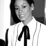 Black and white portrait of a woman with a confident smile, wearing a white top with thin stripes and a black bow tie.