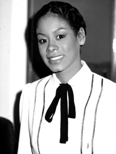 Black and white portrait of a woman with a confident smile, wearing a white top with thin stripes and a black bow tie.