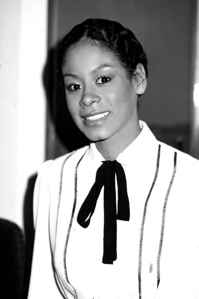 Black and white portrait of a woman with a confident smile, wearing a white top with thin stripes and a black bow tie.