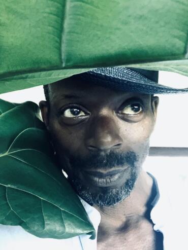 Close-up of a man’s face peeking through large green leaves, with a black hat partially visible.