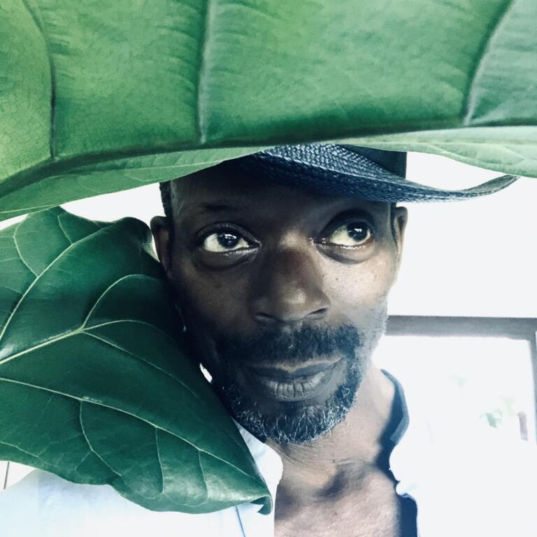Close-up of a man’s face peeking through large green leaves, with a black hat partially visible.