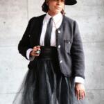 Elegant woman in a black hat, textured jacket, white shirt, and layered tulle skirt, blending classic and contemporary style.