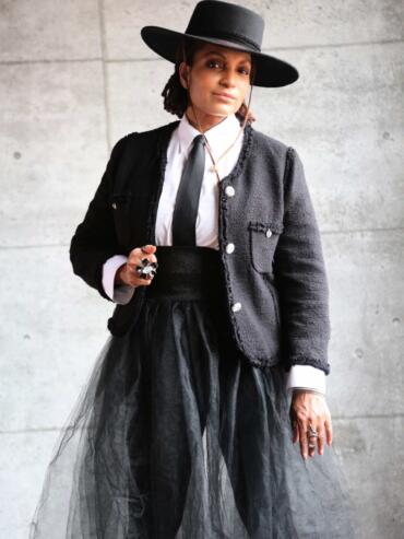 Elegant woman in a black hat, textured jacket, white shirt, and layered tulle skirt, blending classic and contemporary style.