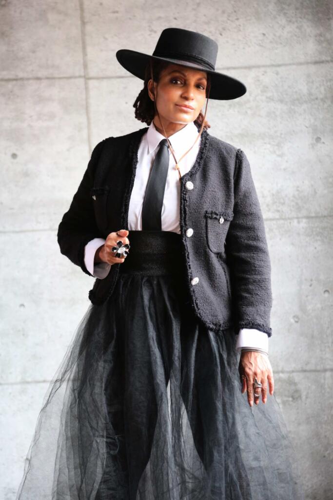 Elegant woman in a black hat, textured jacket, white shirt, and layered tulle skirt, blending classic and contemporary style.