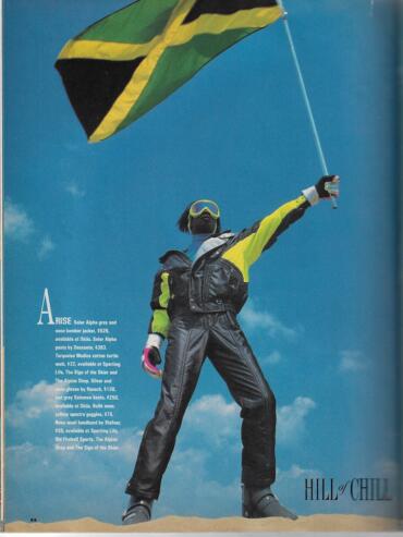 Person standing on sand, waving with a Jamaican flag, dressed in vibrant outdoor gear.