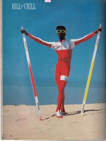 Athlete in a red and white racing suit with sunglasses, holding ski poles on a sandy terrain under a blue sky.
