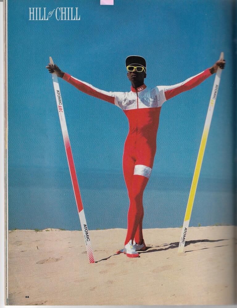 Athlete in a red and white racing suit with sunglasses, holding ski poles on a sandy terrain under a blue sky.