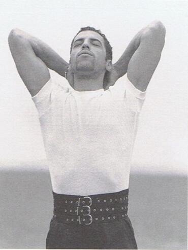 Man with arms behind head, wearing a white t-shirt and a wide belt, against a plain background.