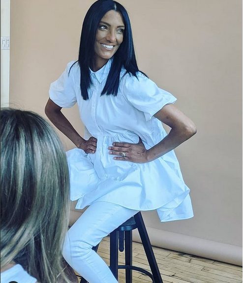 Woman in a white dress sitting on a stool, smiling confidently against a neutral background.