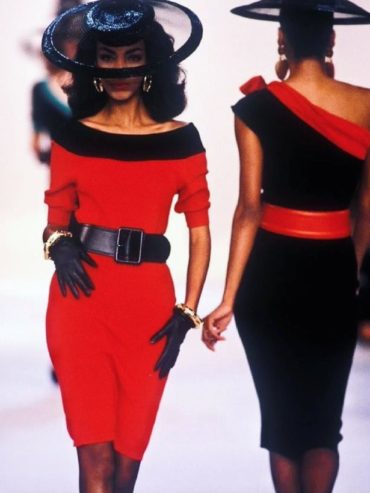 Elegant women in red and black dresses with wide-brim hats walk the runway.