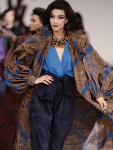 Model walking runway in vibrant patterned coat and navy high-waisted pants at fashion show.